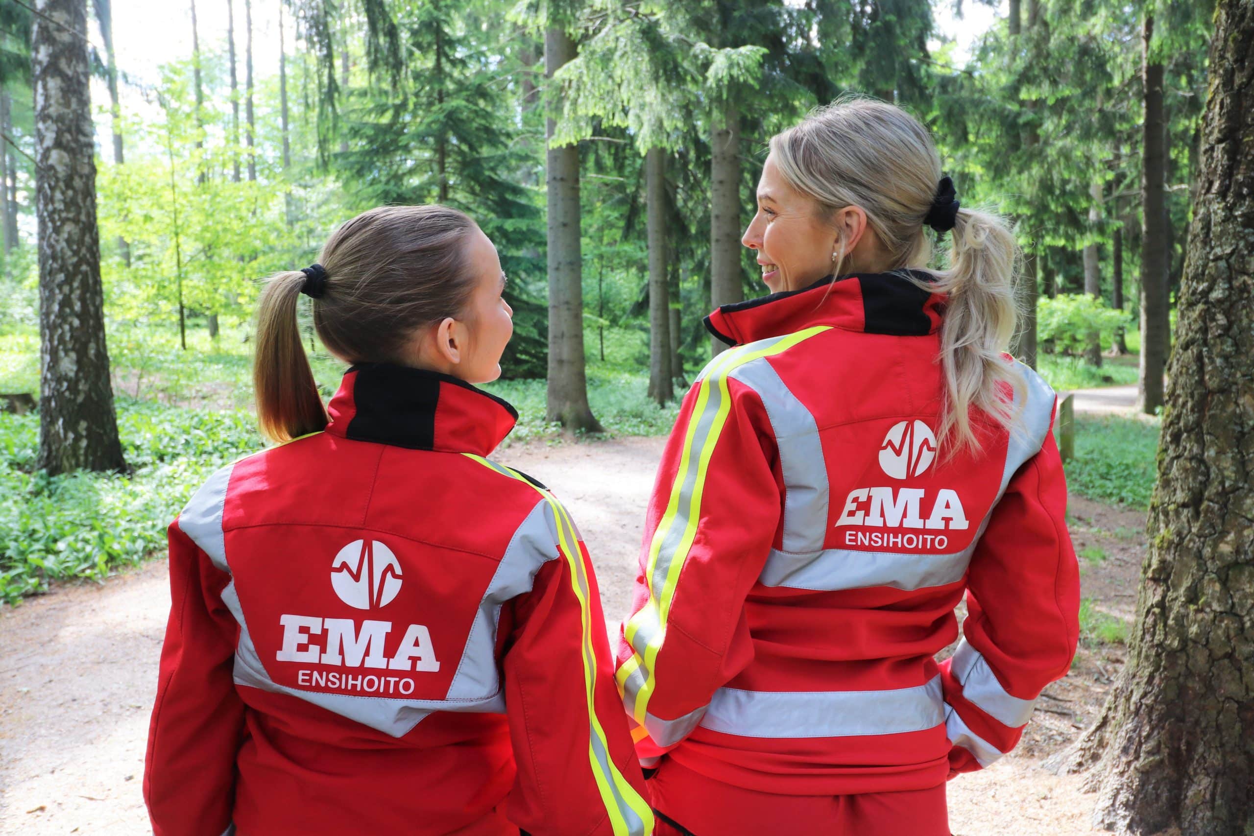 EMA -ensihoidon kaksi työntekijää hymyilee toiselleen katse kamerasta poispäin. Molempien takin selässä näkyy EMA-ensihoidon logo.
